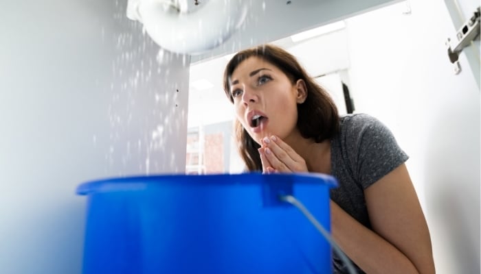 Surprised woman catching water in a bucket from a leaking pipe under the sink