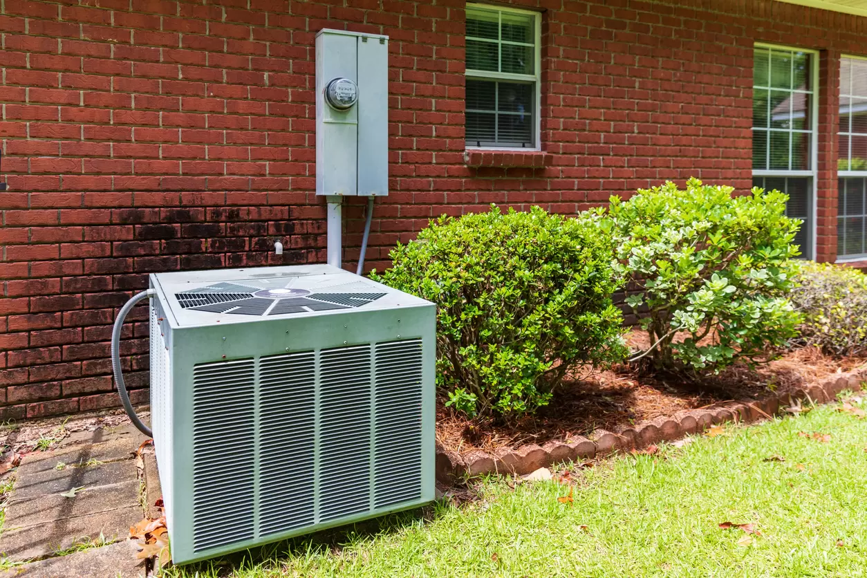 Older style air conditioner system next to home, brick and bushes with clean yard.